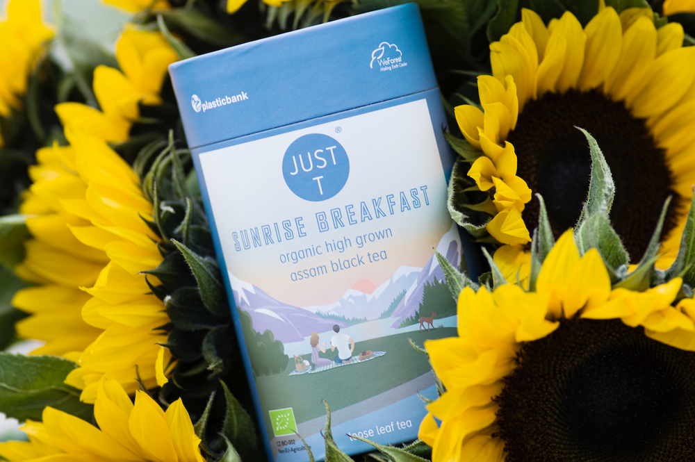 Blaue Teeverpackung "Just T Sunrise Breakfast" inmitten von gelben Sonnenblumen.