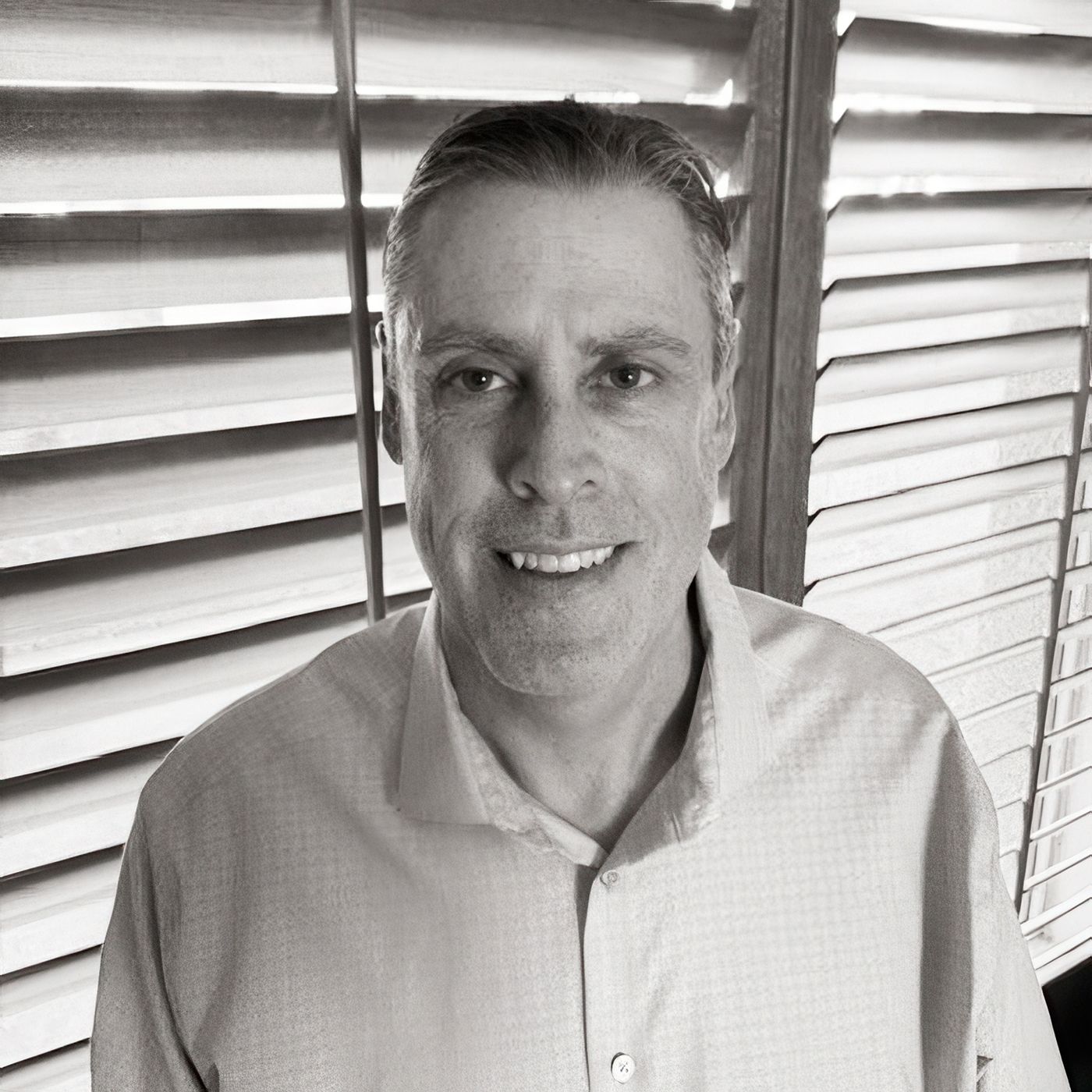 Mann in gestreiftem Hemd steht vor Jalousie, lächelt.