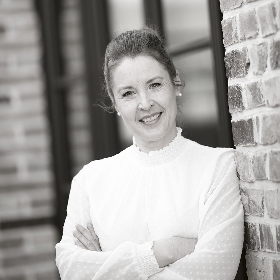 Frau mit hochgestecktem Haar in weißem Oberteil lehnt lächelnd an einer Backsteinwand.