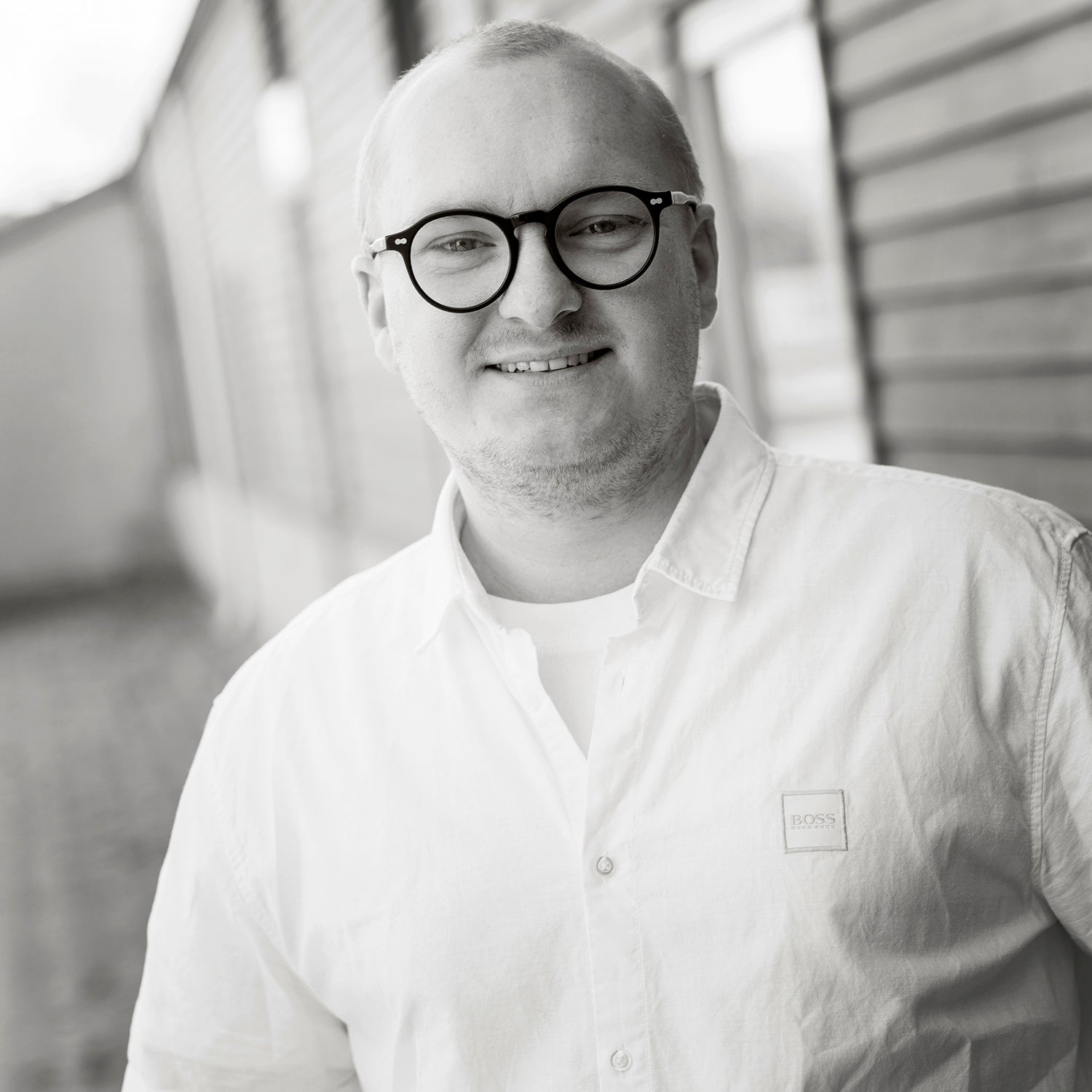 Mann mit Brille und weißem Hemd lächelt in die Kamera, steht vor unscharfem Hintergrund.