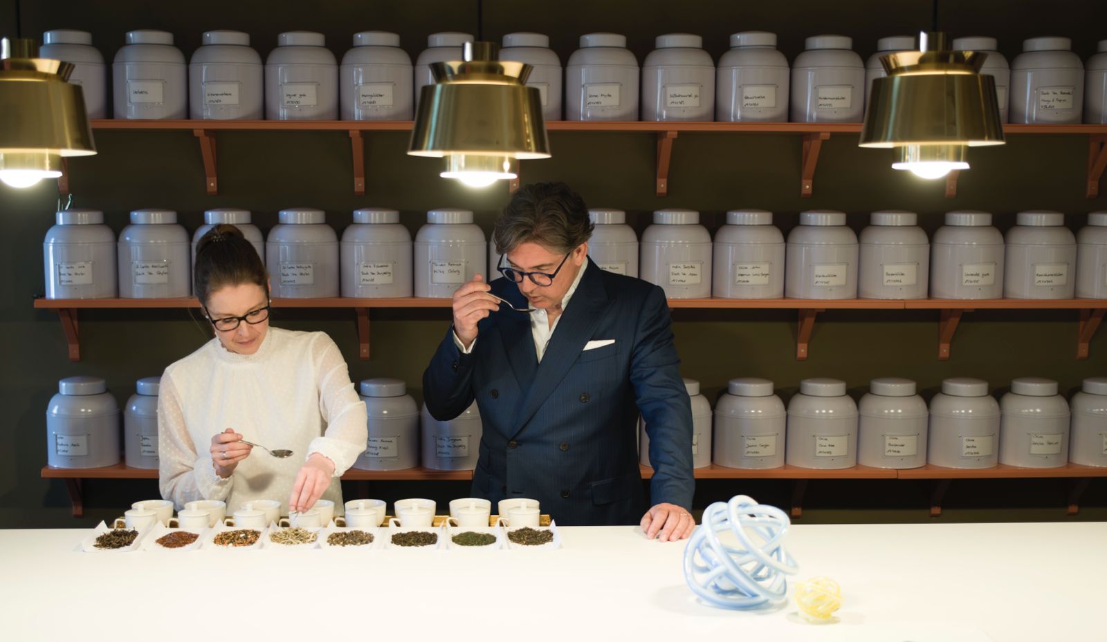 Mann und Frau riechen an Tee, viele Teekannen im Regal dahinter.