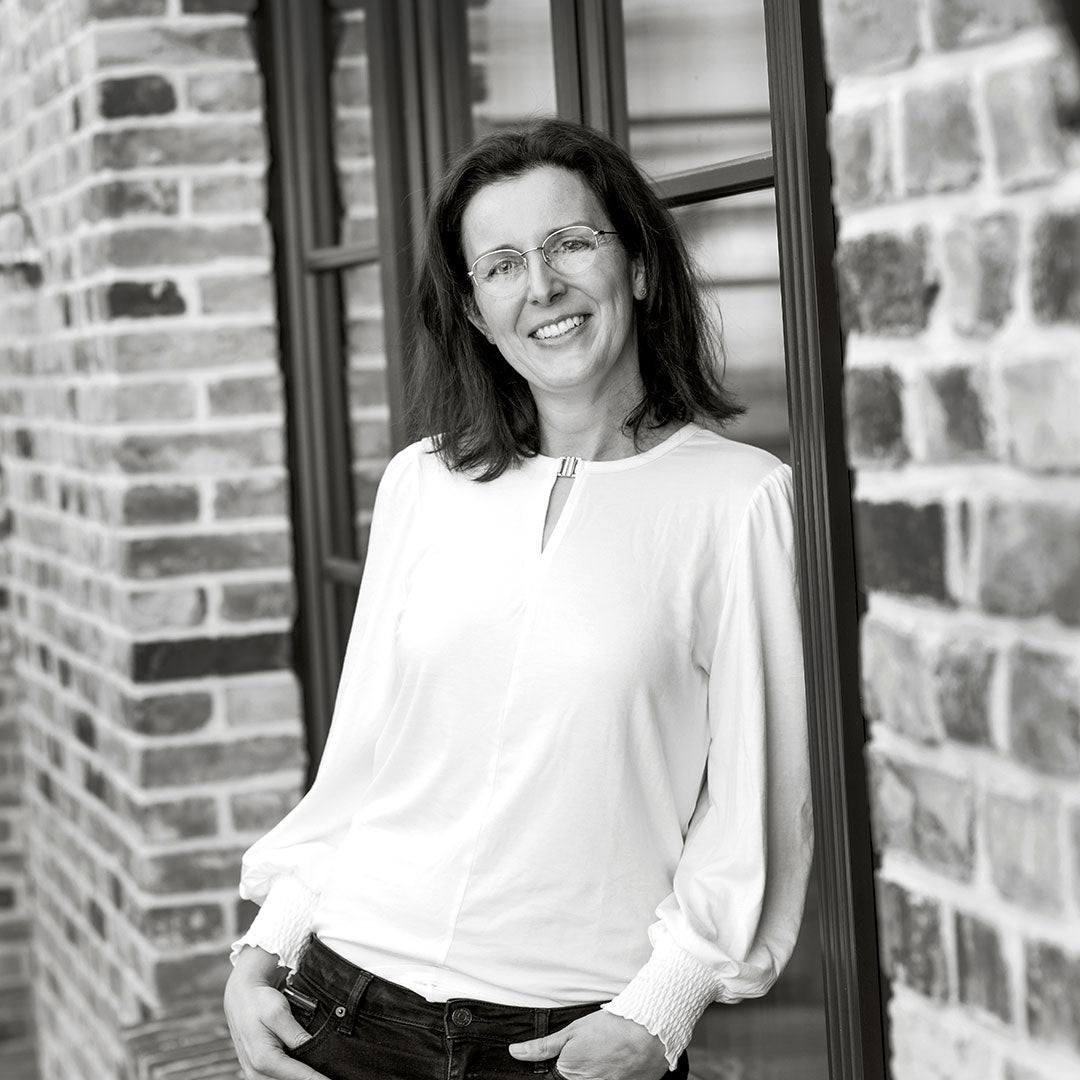 Frau mit Brille und lockigem Haar trägt eine Bluse und lehnt an einer Backsteinfassade.